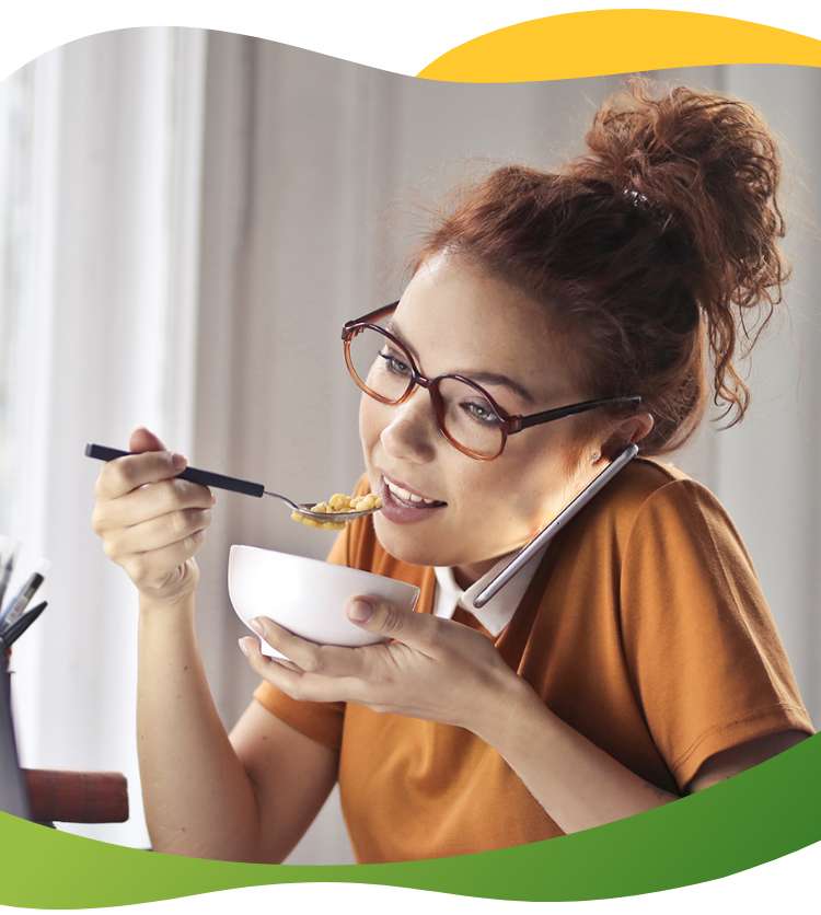Young woman with glasses and high braid sits in front of laptop while talking on phone and eating her meal at the same time